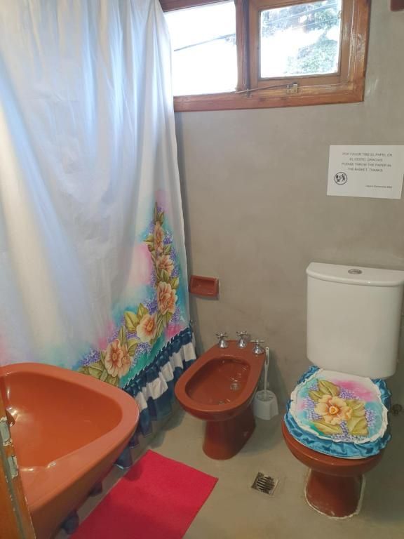 Baño pequeño con lavabo naranja, inodoro y bidé marrones, cortinas azules y fundas para toallas de colores.