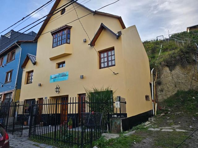 Casa amarilla con ventanas con marcos marrones junto a un edificio azul en una ladera empinada.
