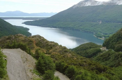 Carretera de montaña con vistas a un lago serpenteante entre verdes colinas y picos lejanos.