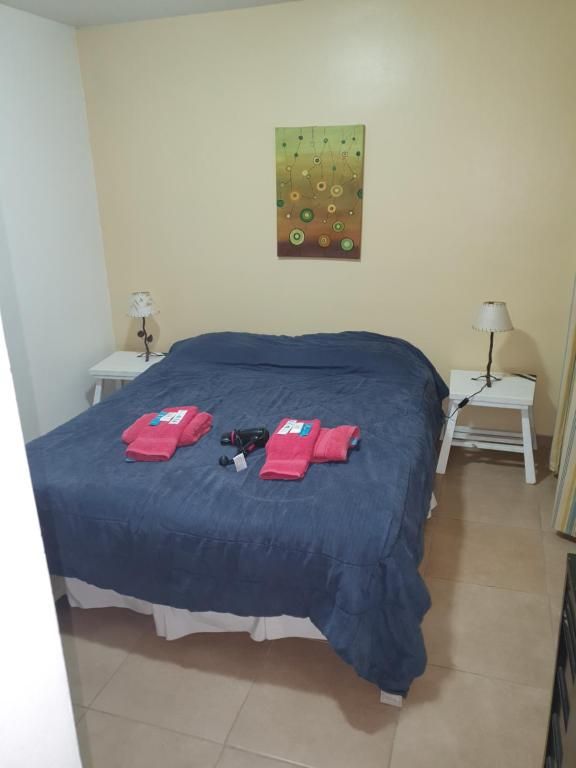 Dormitorio con colcha azul oscuro, dos toallas rosas dobladas y mesitas de noche pequeñas con lámparas.