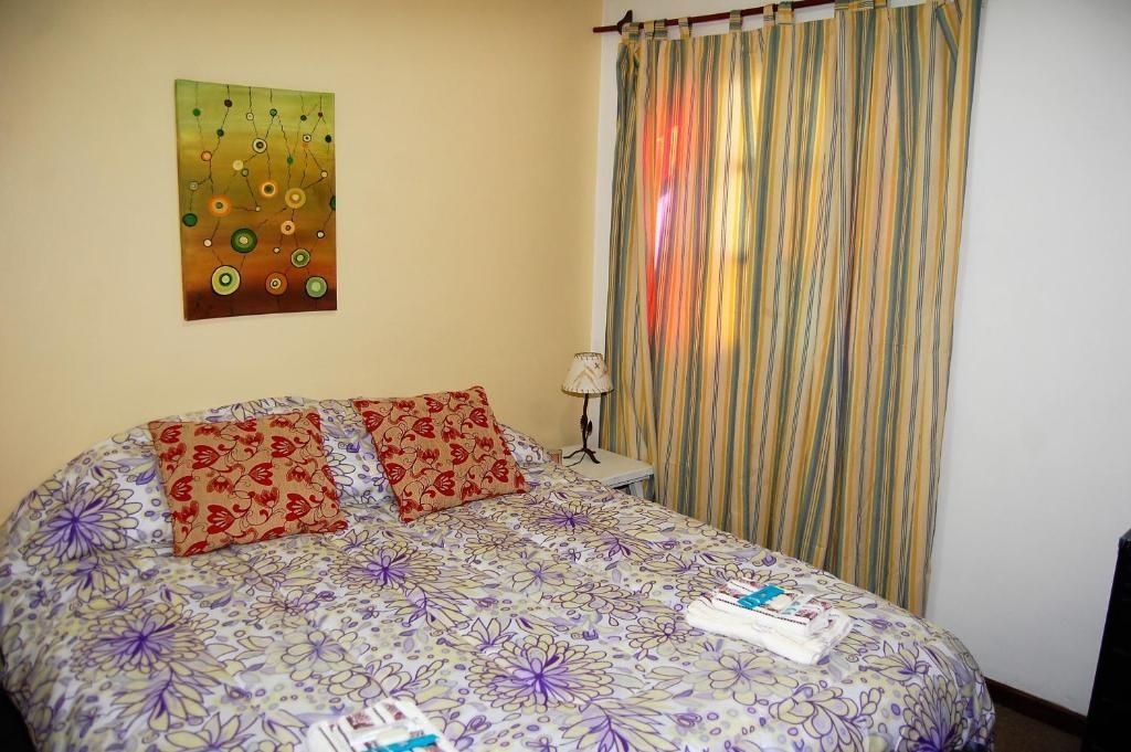 Dormitorio con colcha floral, cojines coloridos, cuadros en la pared y cortinas a rayas junto a la ventana.