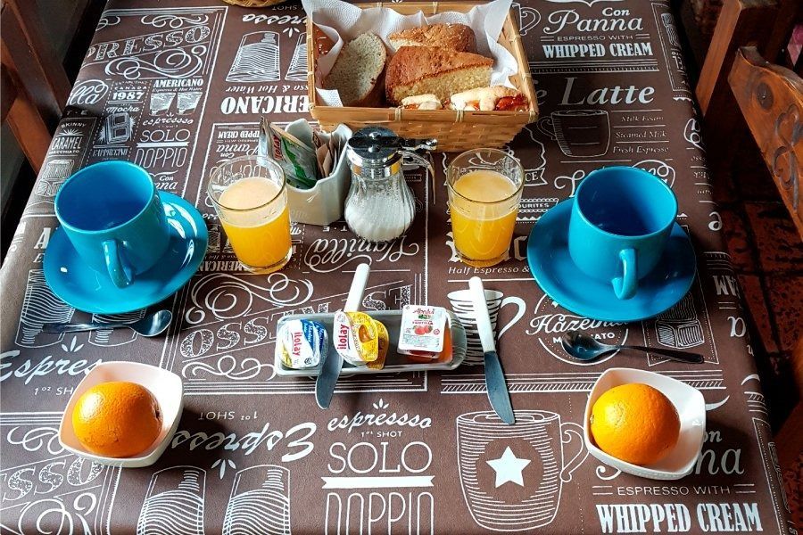 Mesa de desayuno con tazas azules, zumo de naranja, café y pasteles sobre un mantel estampado.