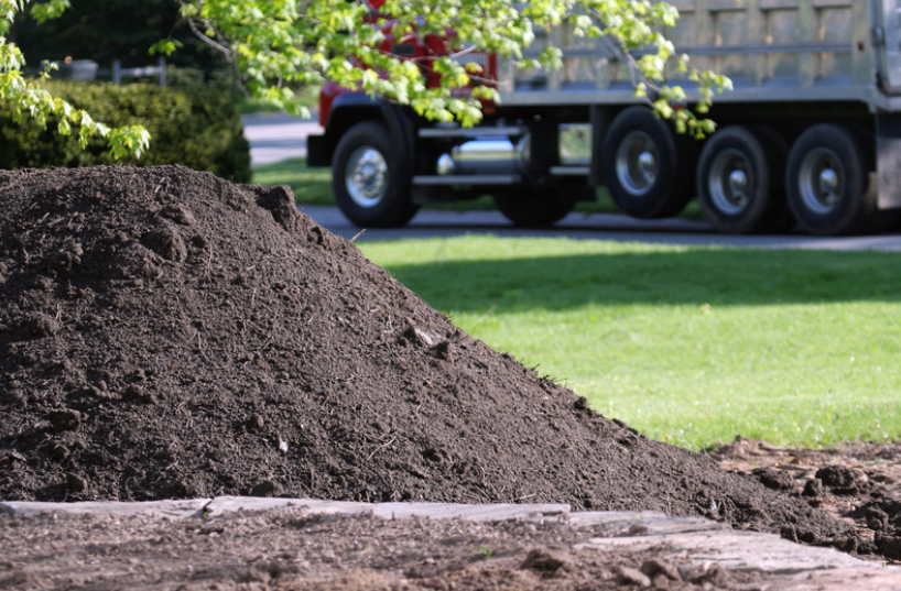 A dump truck is parked in front of a pile of dirt delivered to a driveway