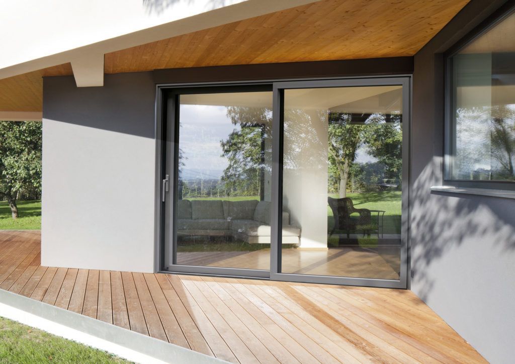 Schuifdeur van glas op een houten dek; grijs frame, reflecterend interieur met uitzicht naar buiten.