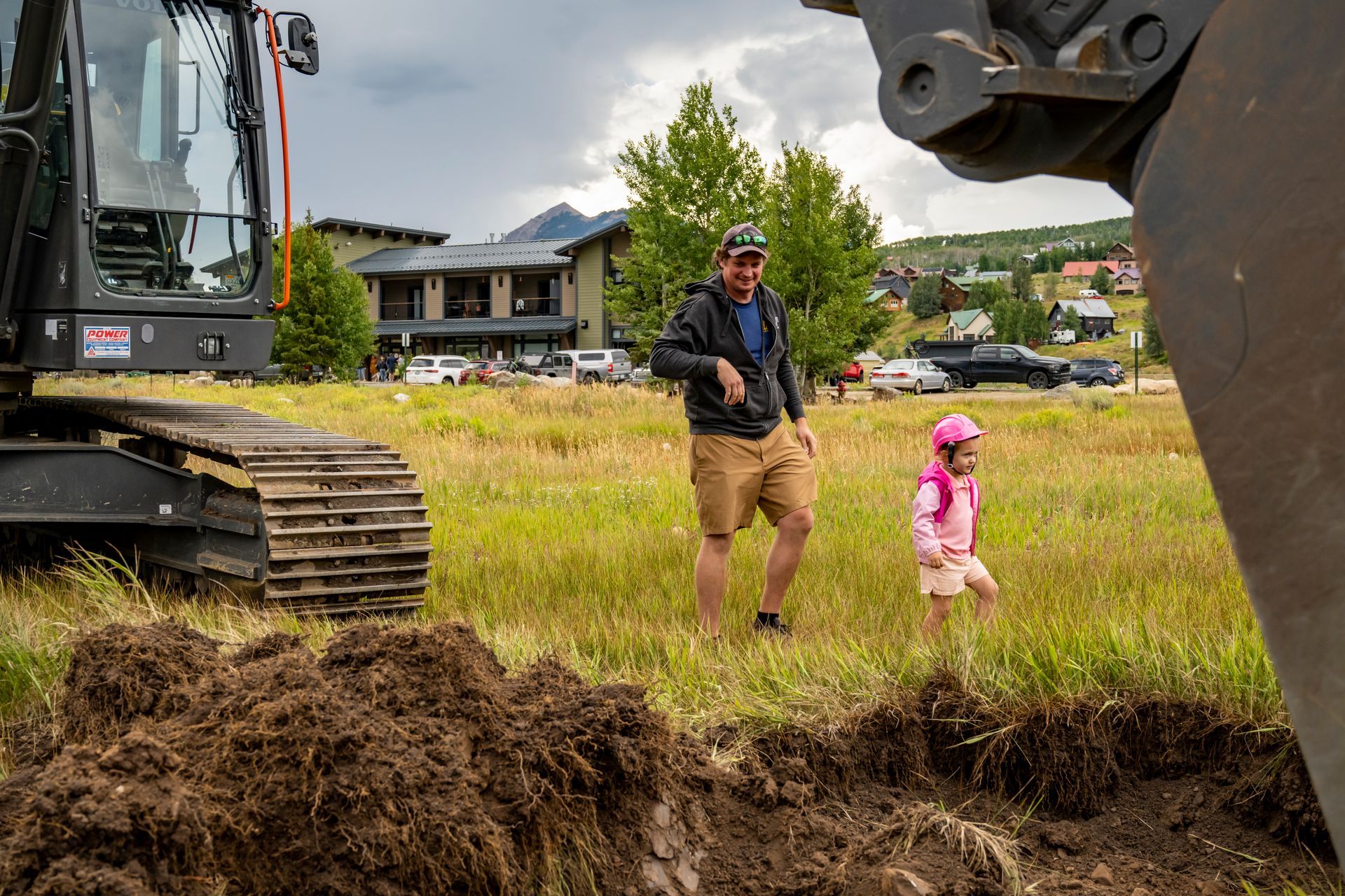 A man and a little girl are standing in a field next to a large excavator.