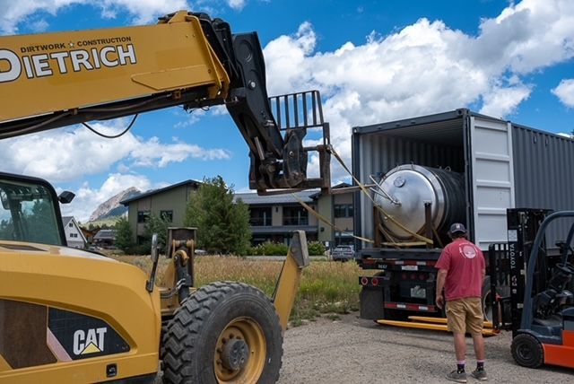 A man in a red shirt is standing next to a forklift and a cat truck.