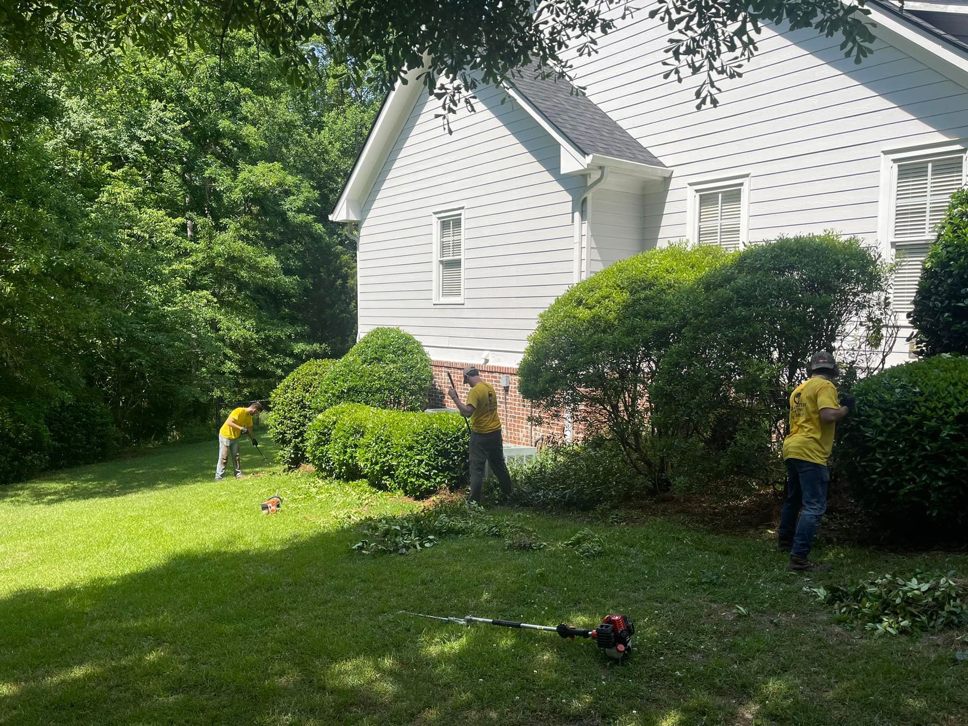 Three workers in yellow shirts trim bushes along the side of a white house on a sunny day.