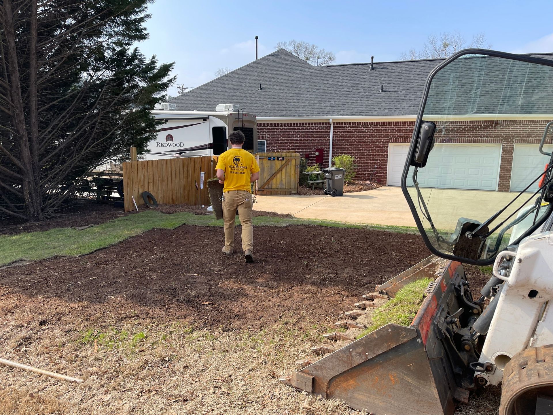 A person in a yellow shirt walks across a yard with fresh mulch, beside a Bobcat loader on a sunny day.