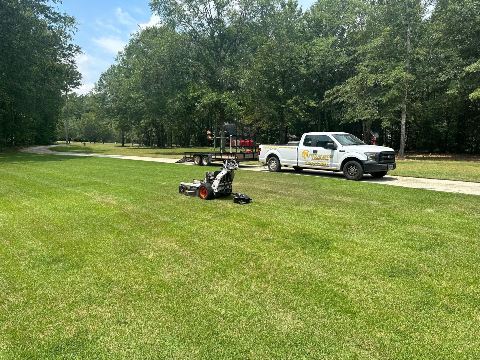 A white work truck with a trailer and a red excavator sits on a grassy field near a treeline.