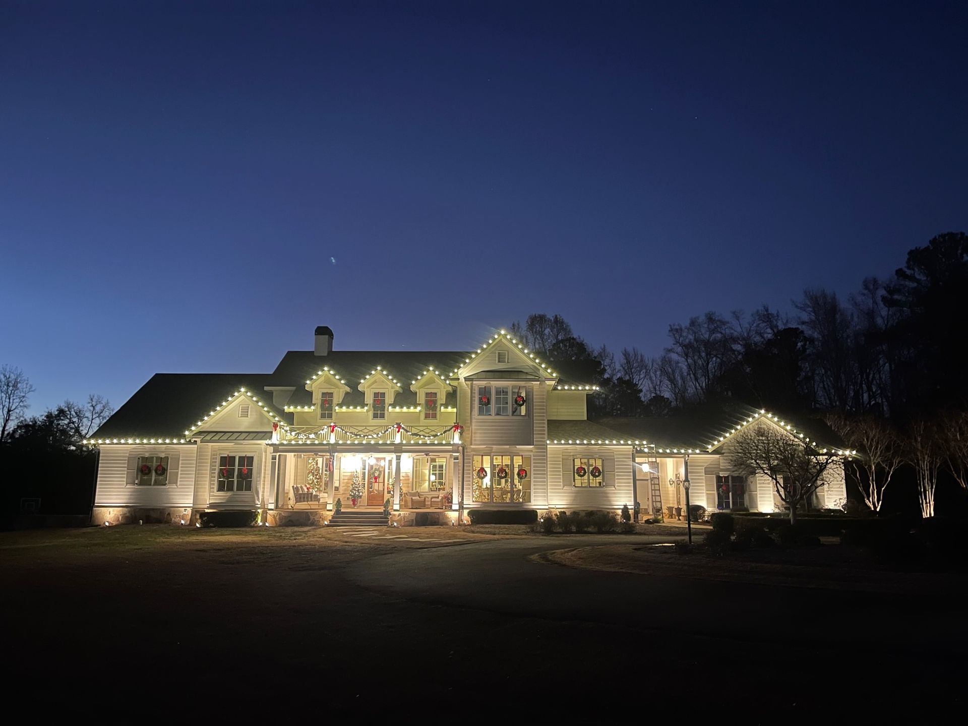 A large house at night, illuminated by warm string lights outlining the roofline and porch against a dark sky.