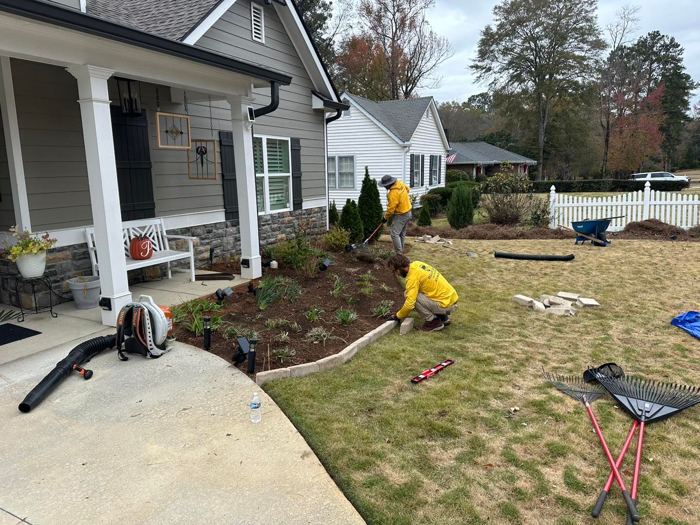 Two workers in yellow shirts landscape a front garden with mulch and stone edging outside a gray house.