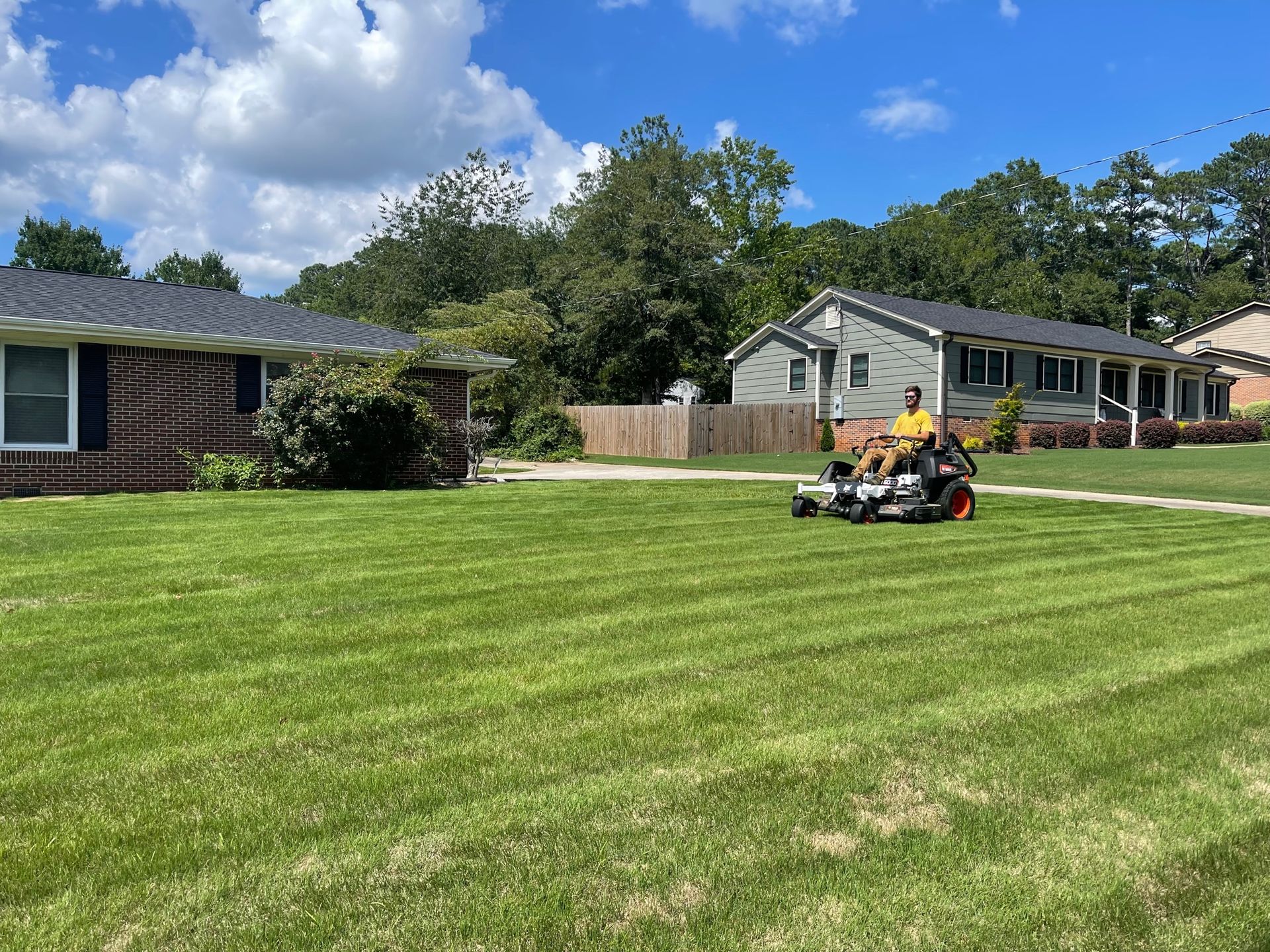 A person rides a zero-turn lawn mower across a large, sunny, mowed residential lawn near two brick and siding houses.