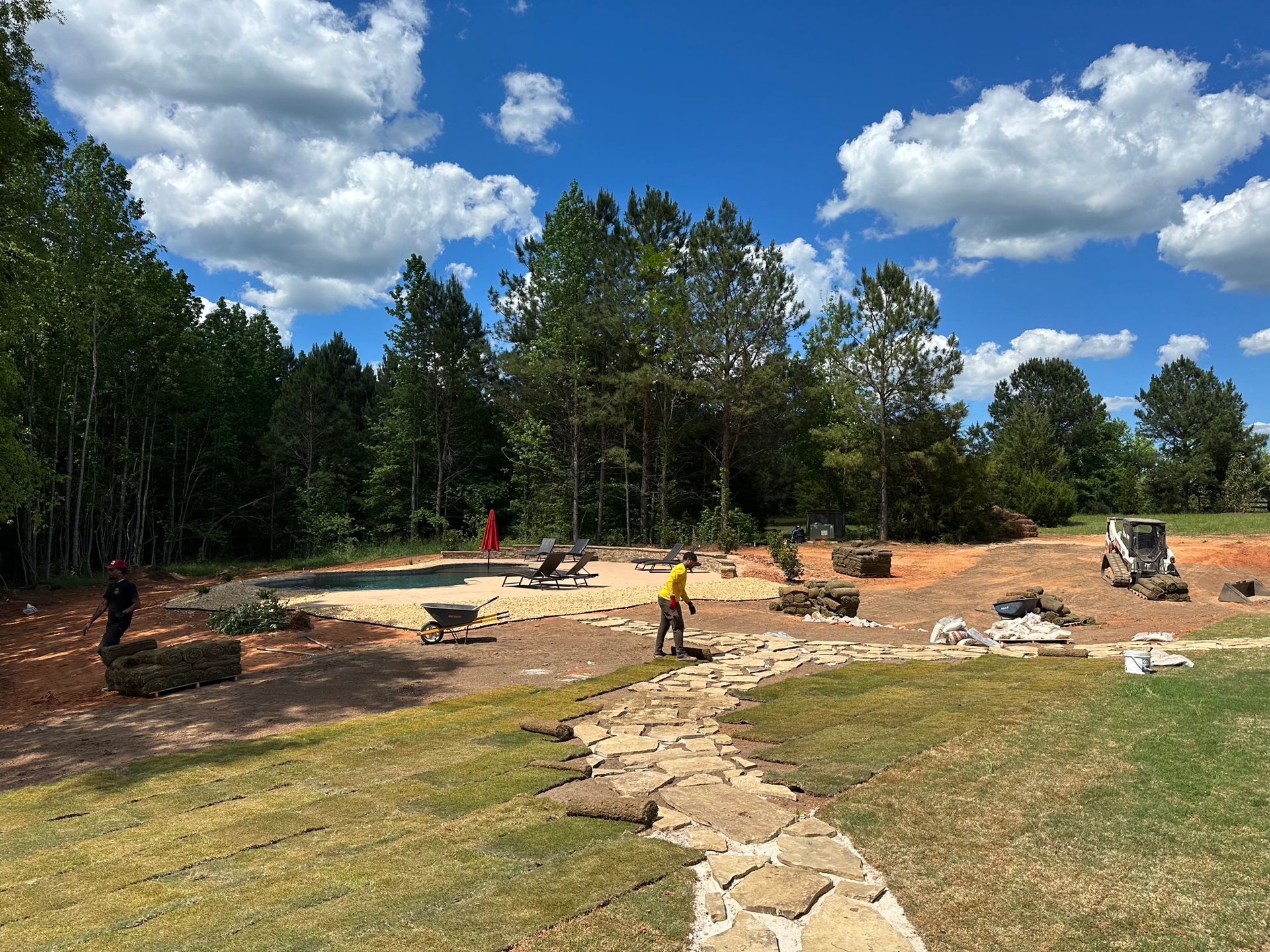 Workers landscape a yard with a stone path, sand pit, and construction machinery under a sunny sky with scattered clouds.