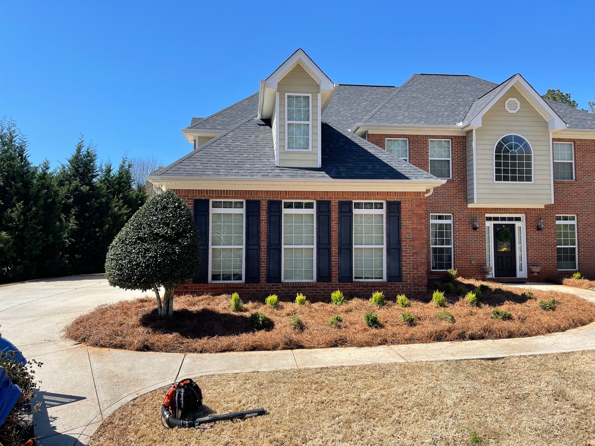 A two-story brick and siding house with a mulched garden bed and a leaf blower on the lawn under a clear blue sky.