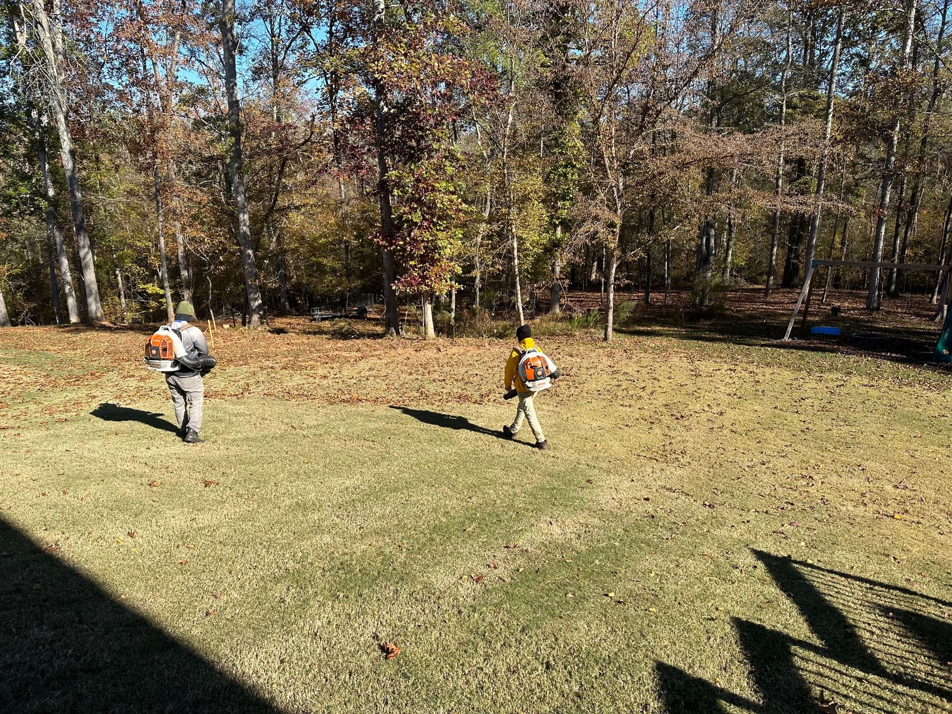 Two workers with leaf blowers clear dry leaves from a grassy lawn in front of a wooded area.