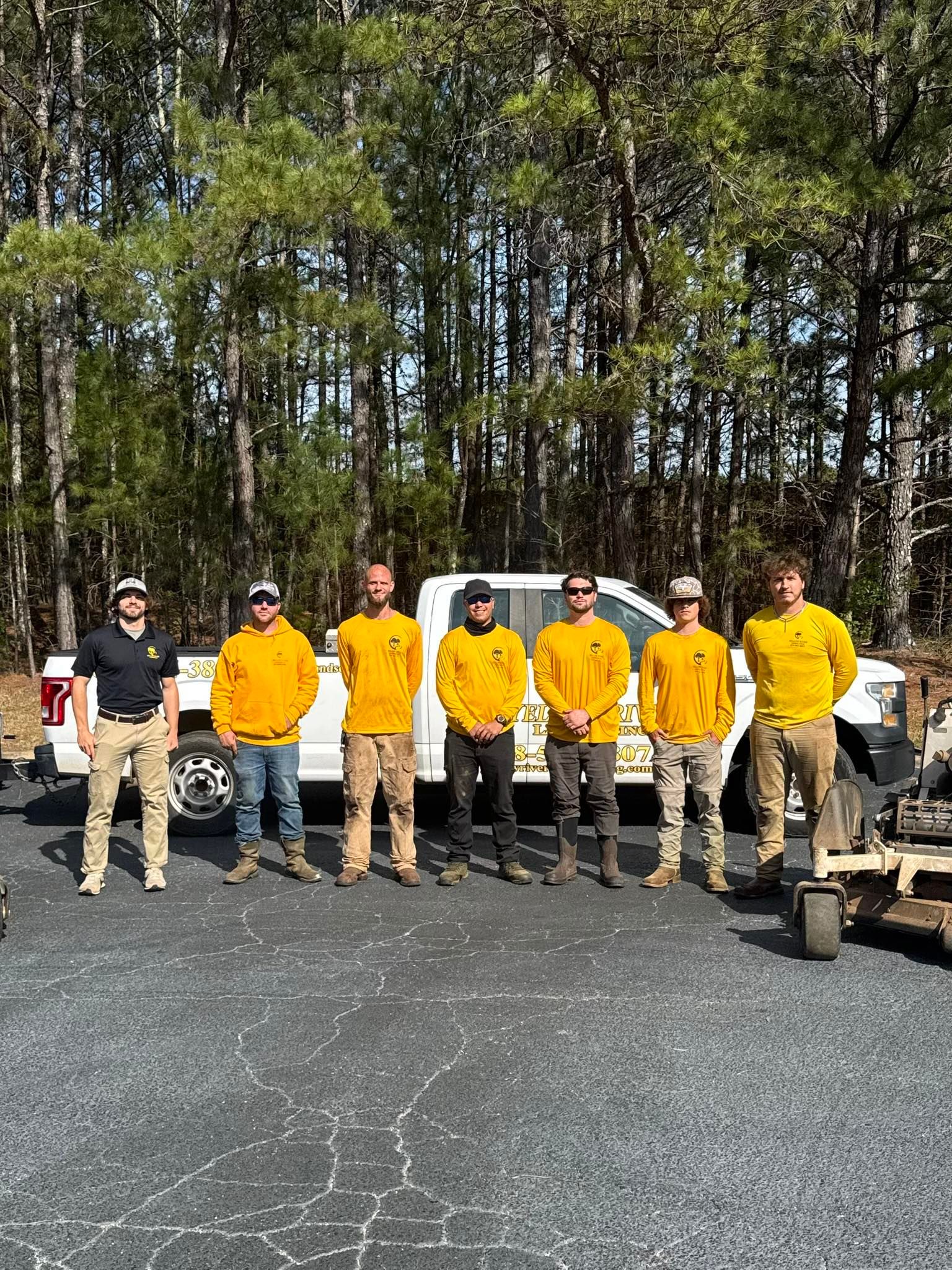 A group of seven people in matching yellow work shirts standing in a row in front of a white truck in a wooded area.