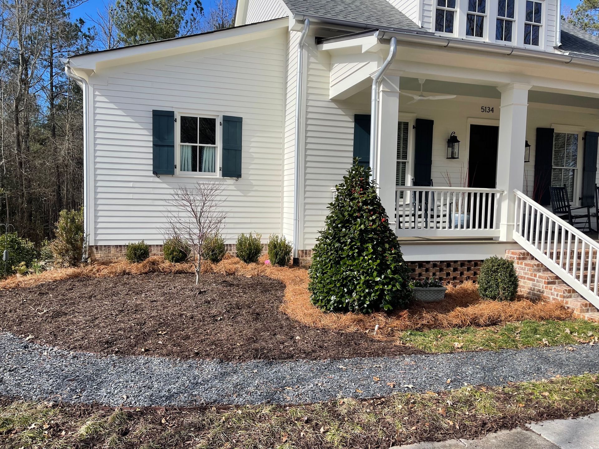 A white house with black shutters, a front porch, and a landscaped yard with pine straw mulch and a gray stone path.