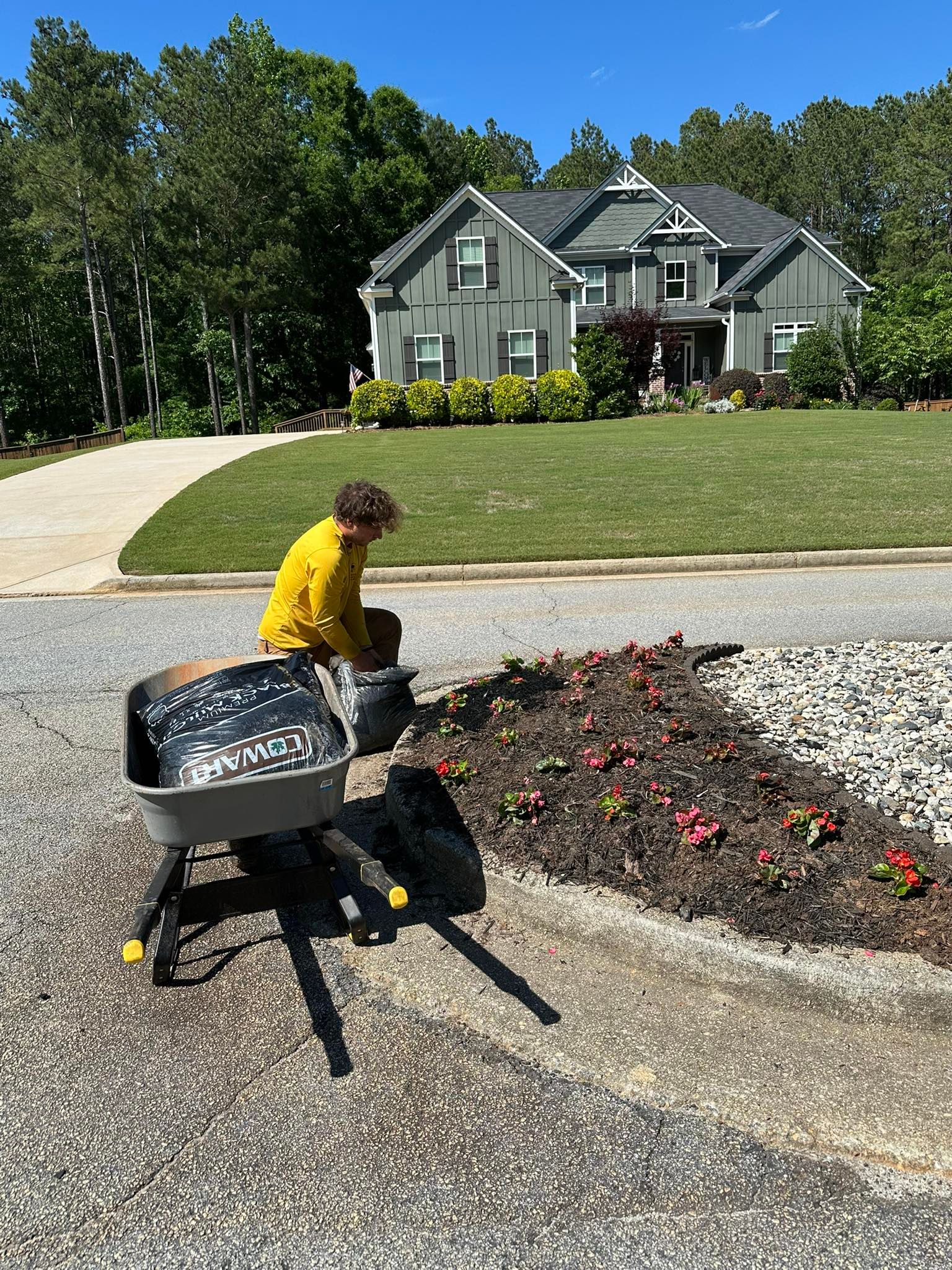 A person in a yellow shirt kneels in a driveway next to a wheelbarrow, planting flowers in a garden bed near a house.