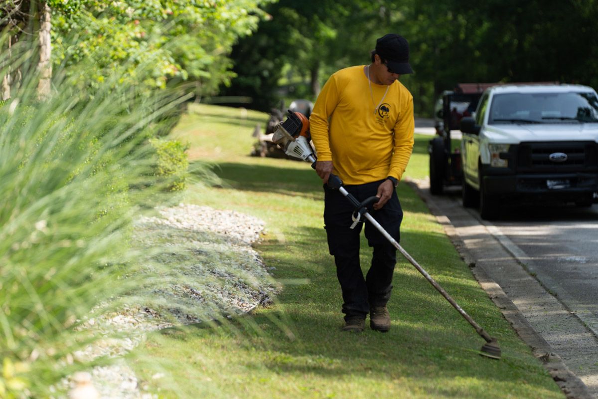 A worker in a yellow long-sleeved shirt uses a string trimmer to edge a lawn along a road next to a white pickup truck.