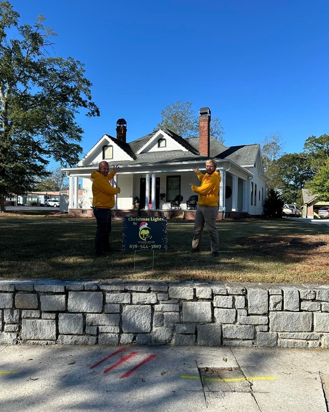 Two people in yellow jackets stand in front of a white house with a stone wall in the foreground on a sunny day.