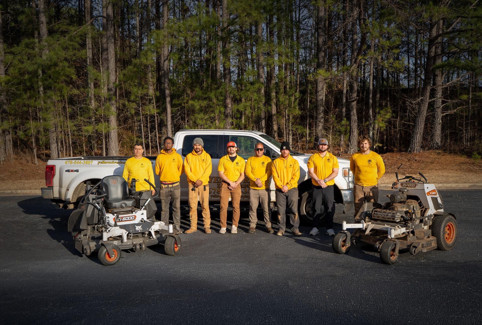 A group of people in yellow uniforms standing in front of a white pickup truck with two commercial lawn mowers.