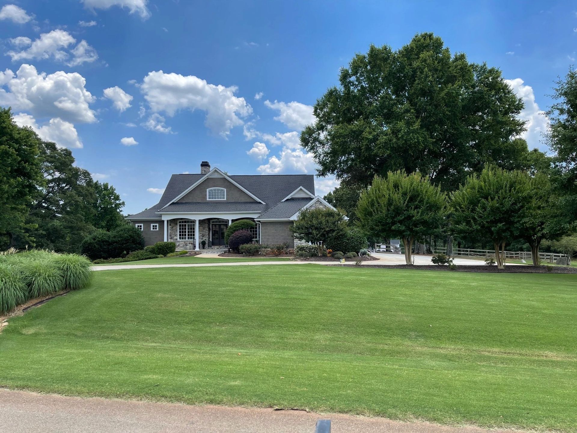 A gray house with a prominent front porch and roof gables, surrounded by a lush green lawn and mature trees under a blue sky.