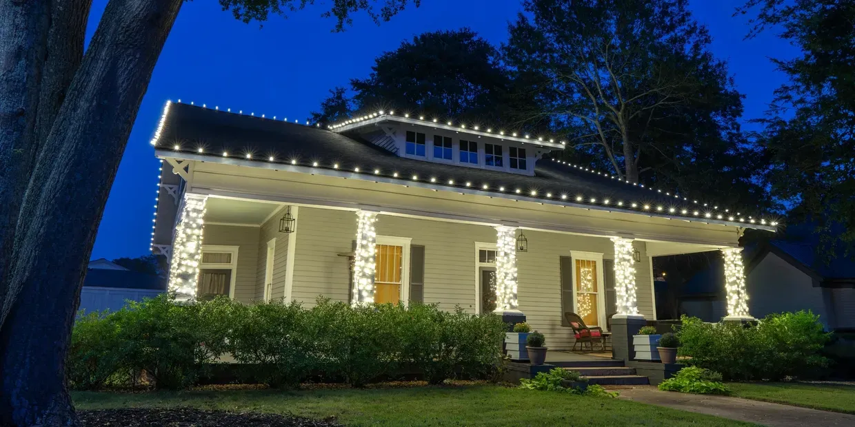 A house at dusk decorated with warm white string lights wrapped around the porch pillars and roofline.
