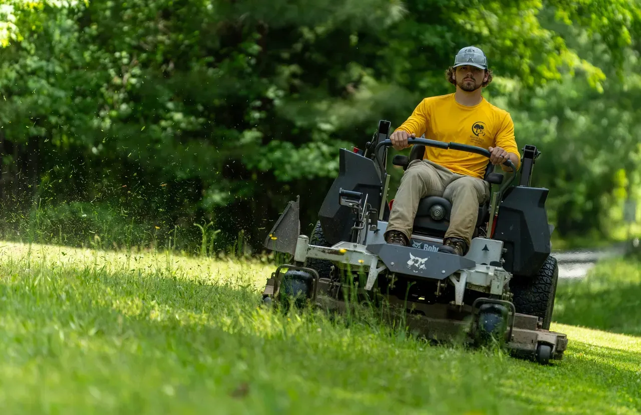 A person wearing a yellow shirt and a cap drives a silver Bobcat zero-turn mower across a sunny, grass-covered hillside.