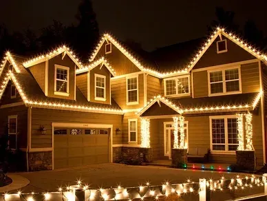 A two-story house at night, decorated with white holiday lights lining the roof, porch columns, and yard fence.