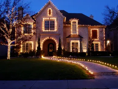 A large, multi-story brick house at dusk, decorated with warm, glowing string lights outlining the roof and windows.