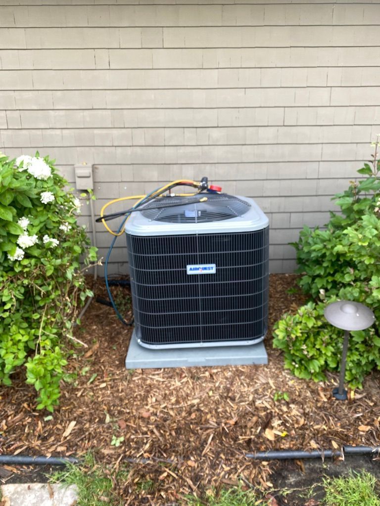 Outdoor air conditioner unit beside a brick wall, with hoses on top and shrubs around it