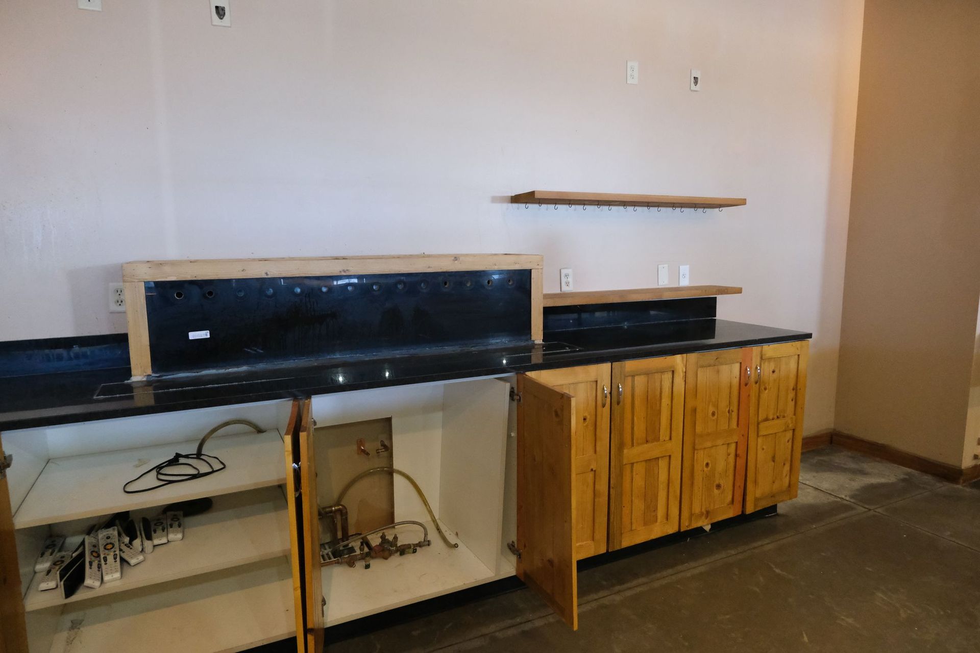 Open wooden cabinets under black countertop bar with exposed plumbing.