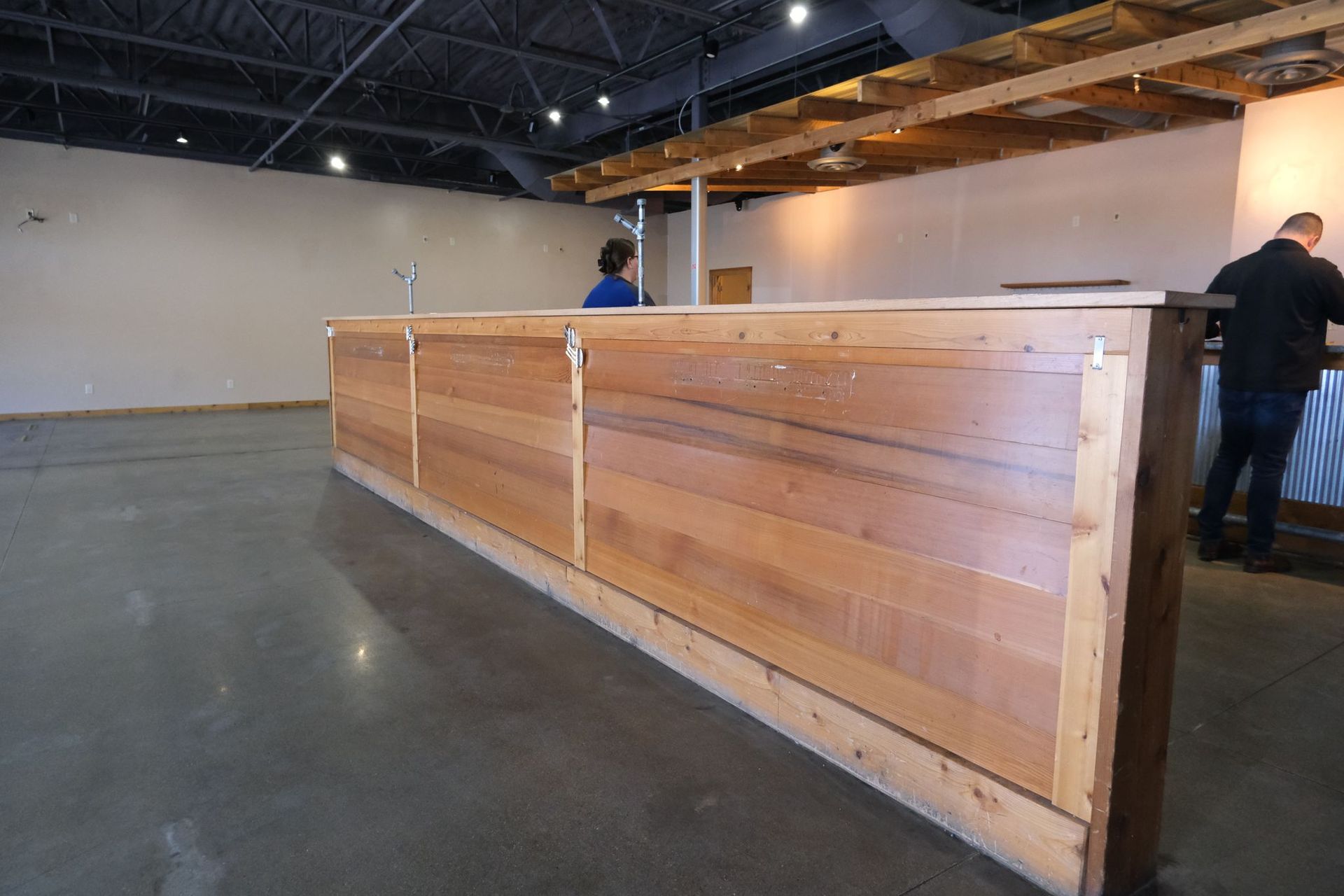 A long, wooden counter with two people standing near it inside a building with a concrete floor.