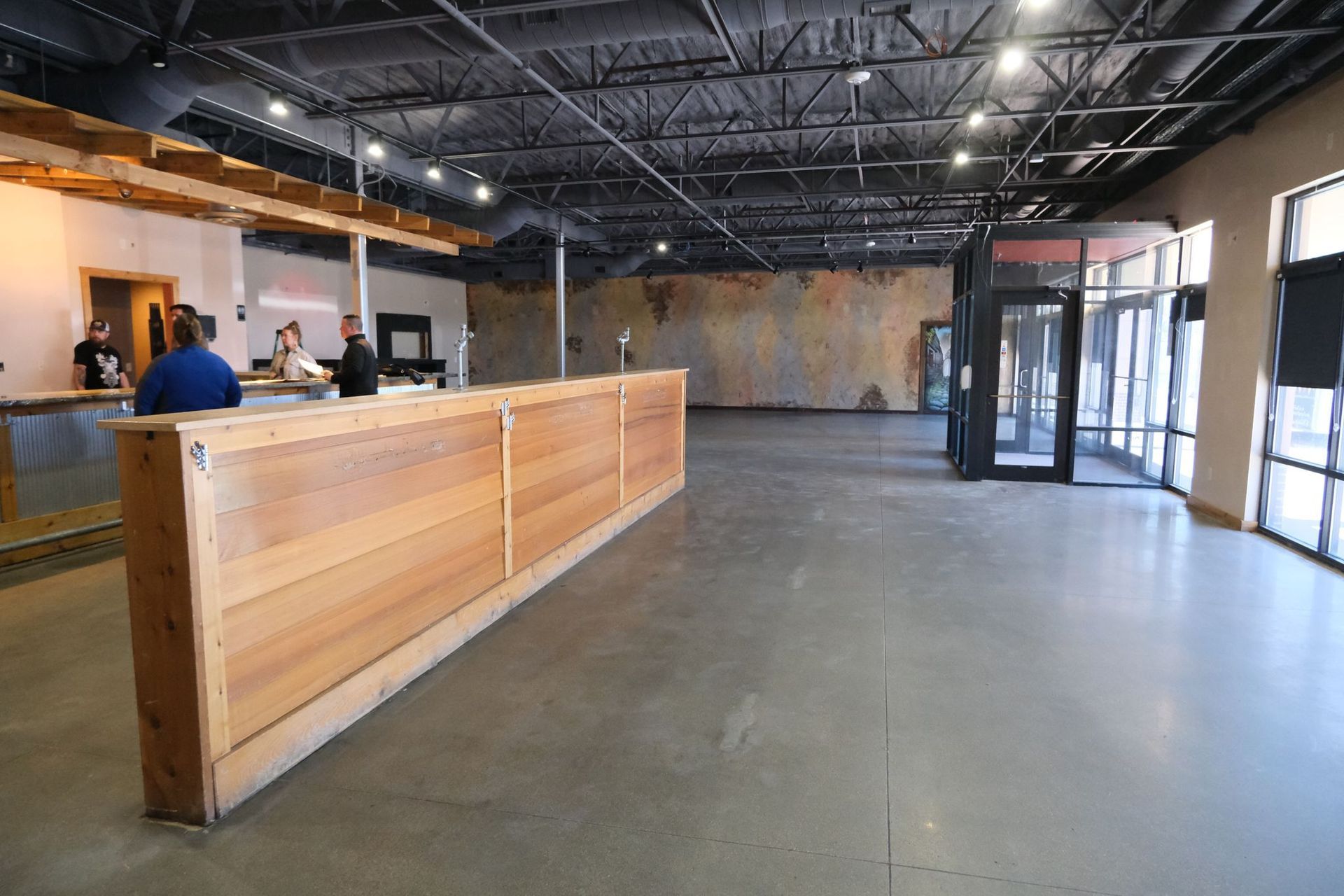 Empty brewery space with a wooden bar, concrete floor, and glass entrance.
