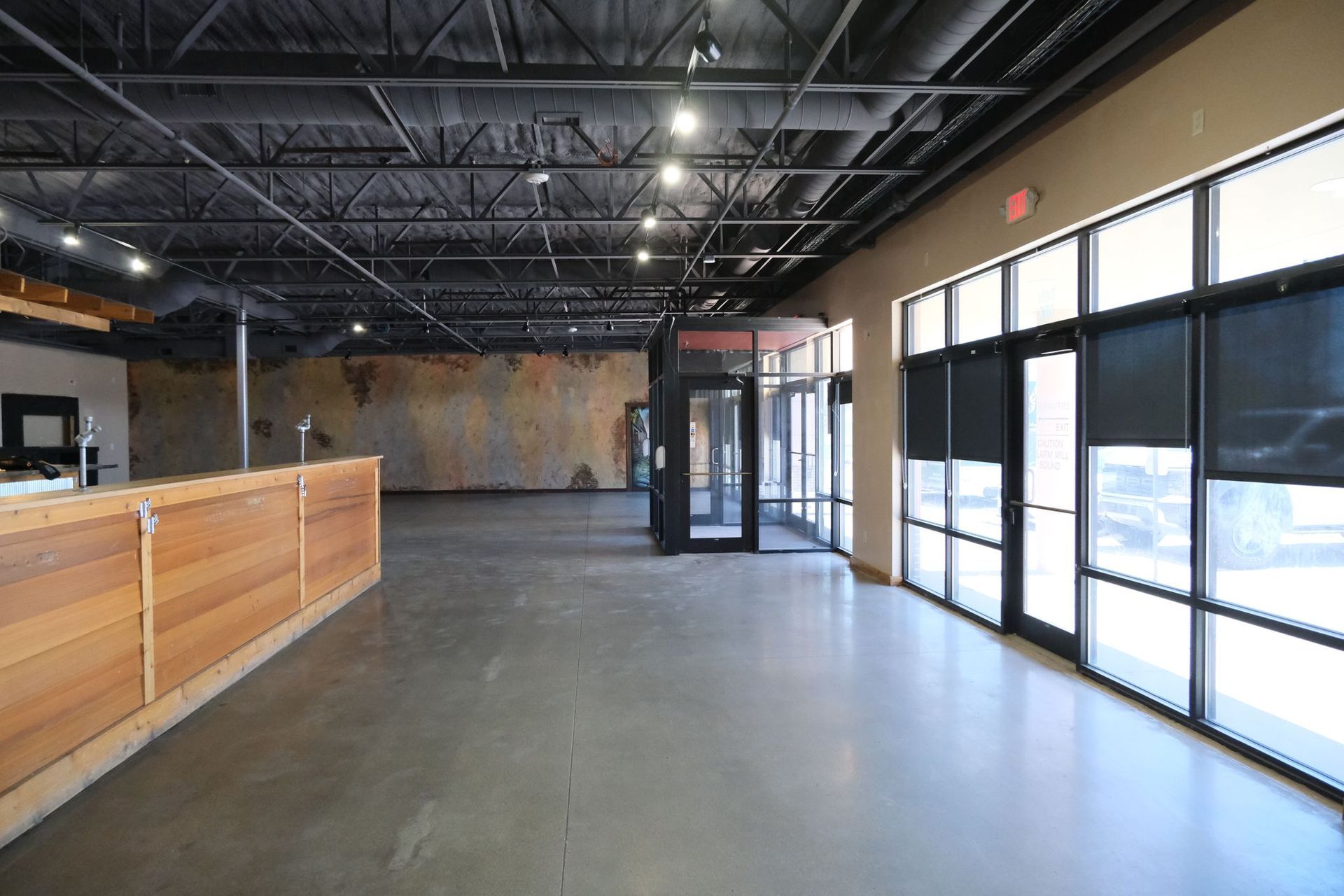 Empty commercial space with a concrete floor, large windows, and a wooden bar.