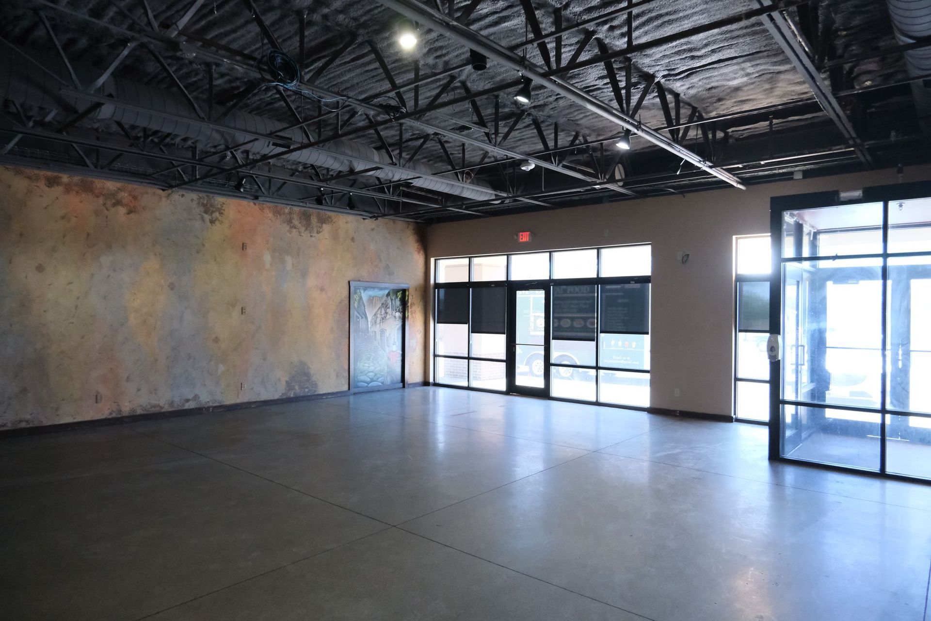 Empty commercial space with a concrete floor and exposed ceiling. Large windows and a colorful wall.