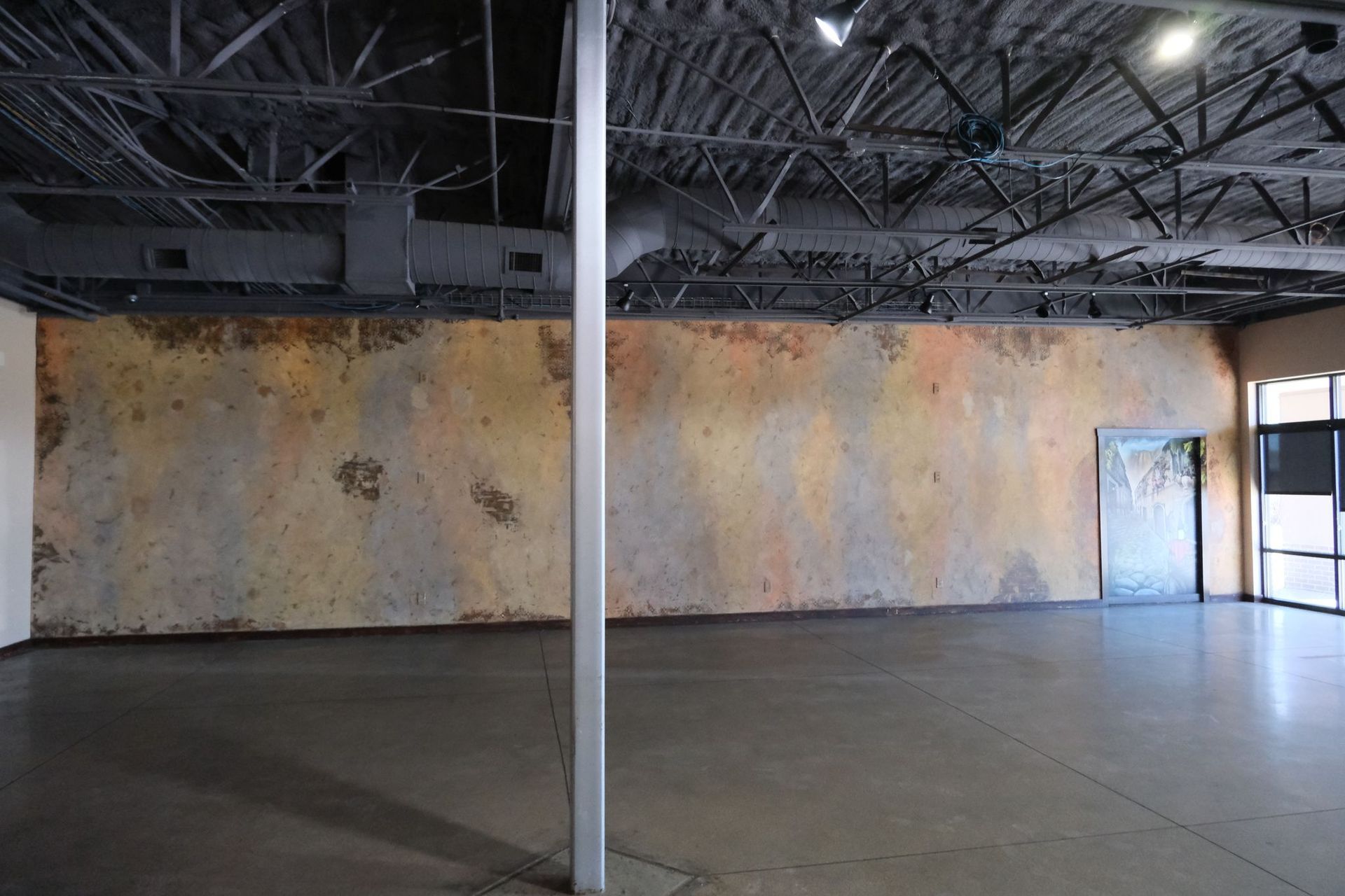 Empty industrial space with concrete floor, high ceiling, and weathered tan wall.