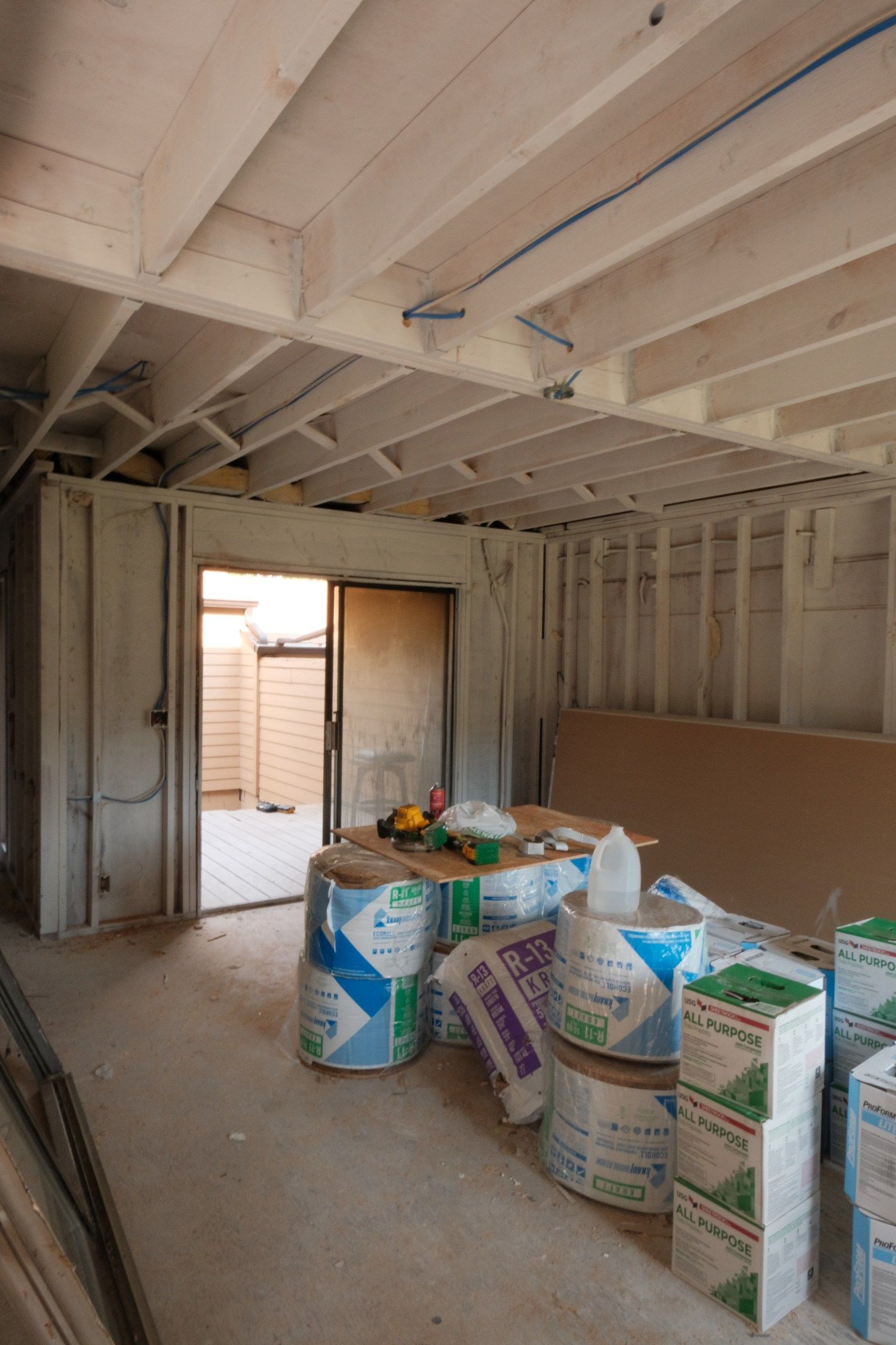 Interior of a room under construction. Insulation rolls and drywall are piled up. Open doorway leads outside.
