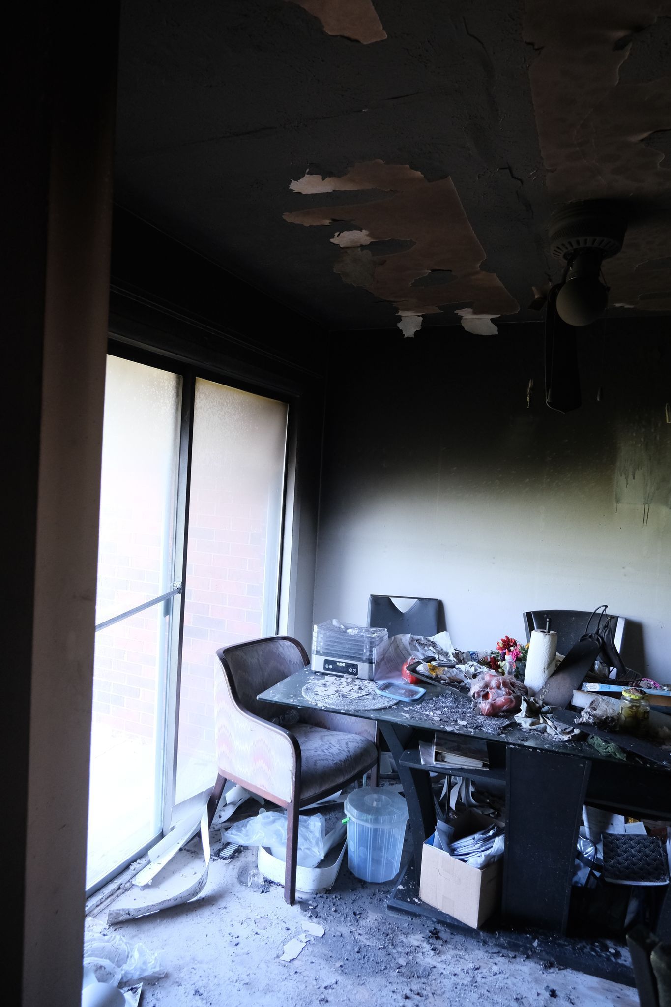 Interior of a room damaged by fire; charred walls and ceiling; table covered with debris and a damaged chair.