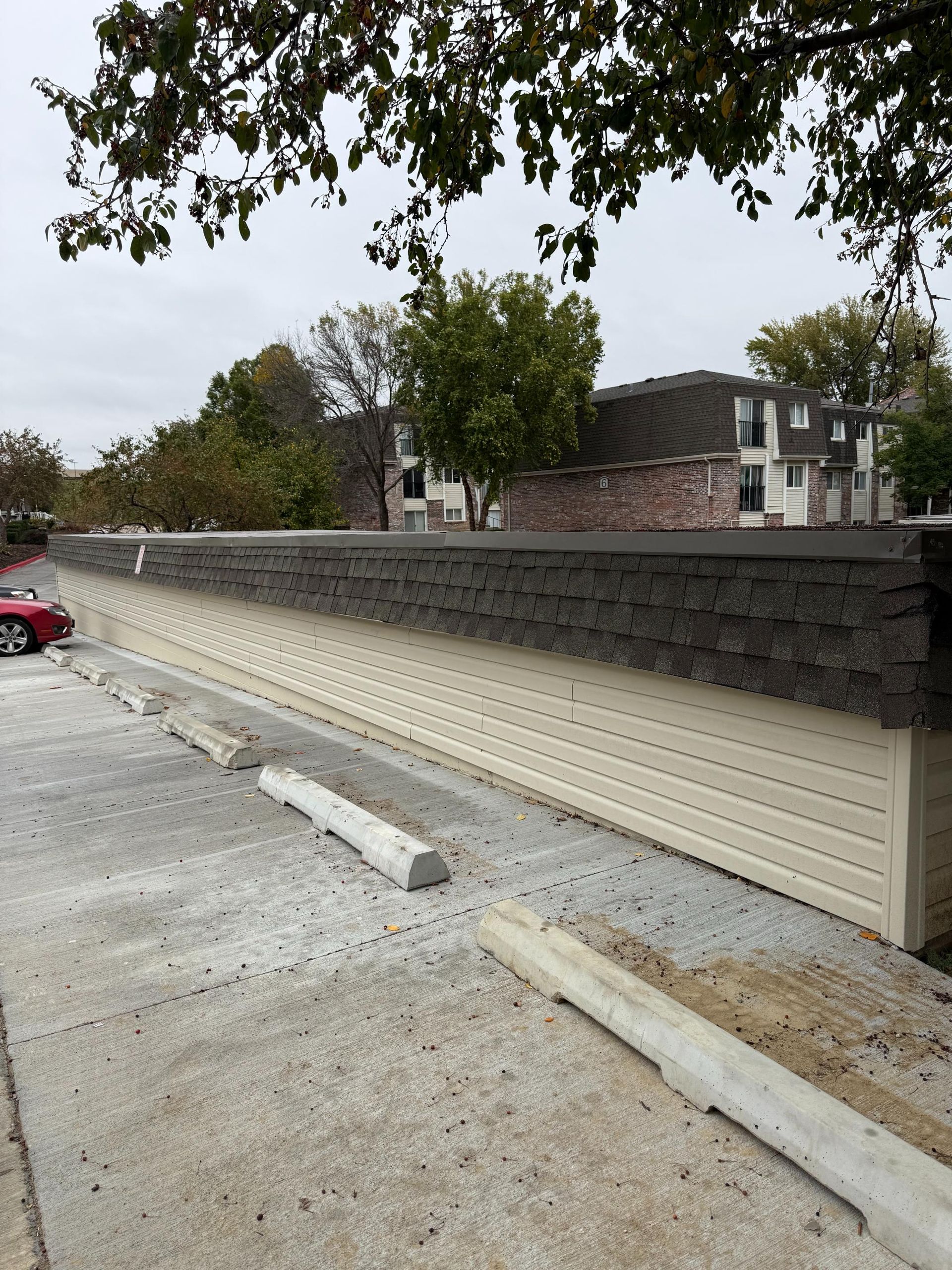 Concrete parking lot with tan-colored siding wall topped with dark gray shingles. Buildings and trees in background.