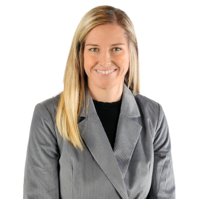 A headshot of a person wearing a light blue button-down shirt and a charcoal blazer against a plain gray background.