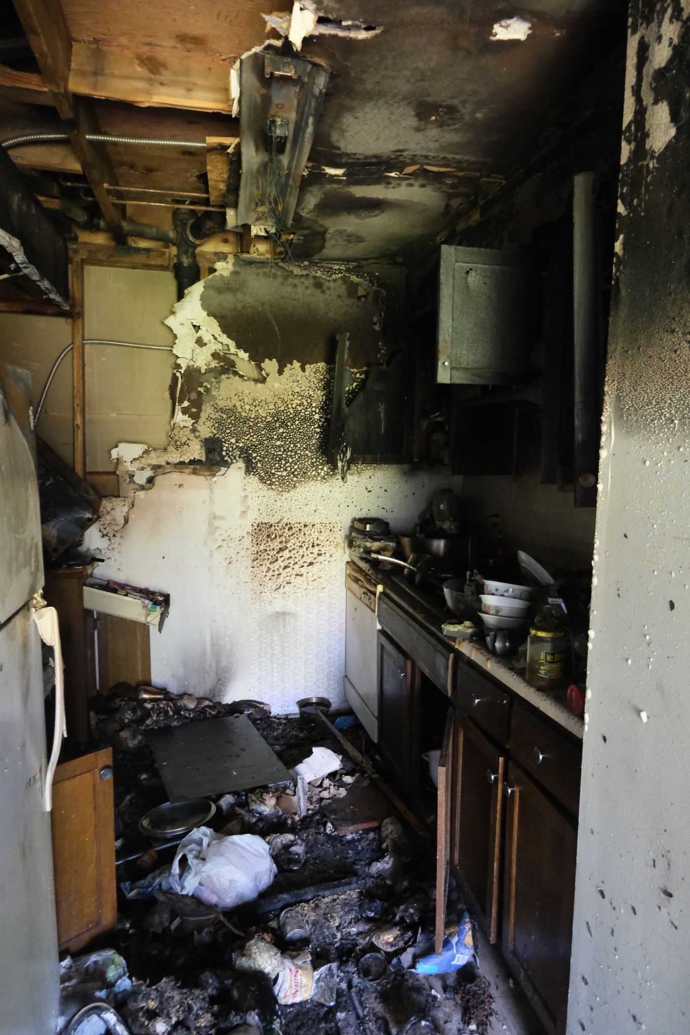Fire-damaged kitchen interior. Charred debris covers floor, walls are blackened and cabinets are visible.