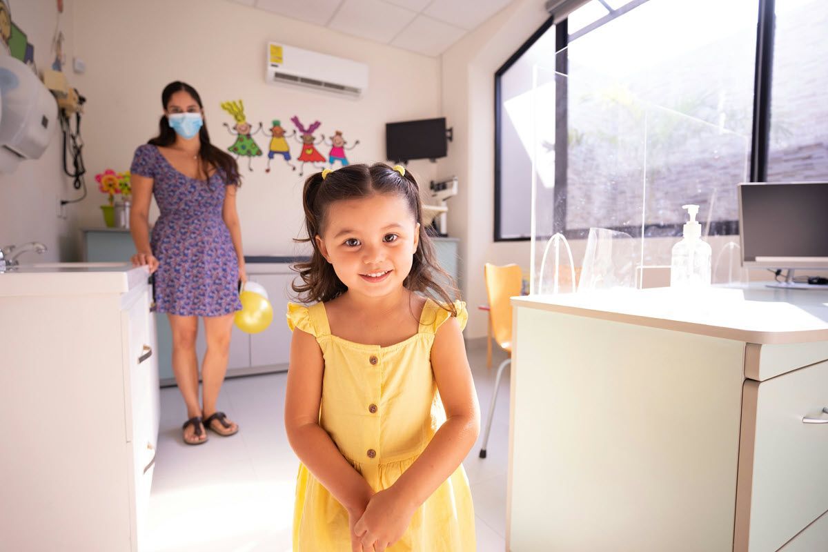 Smiling child in a yellow dress stands in a bright clinic waiting room with a masked woman in the background