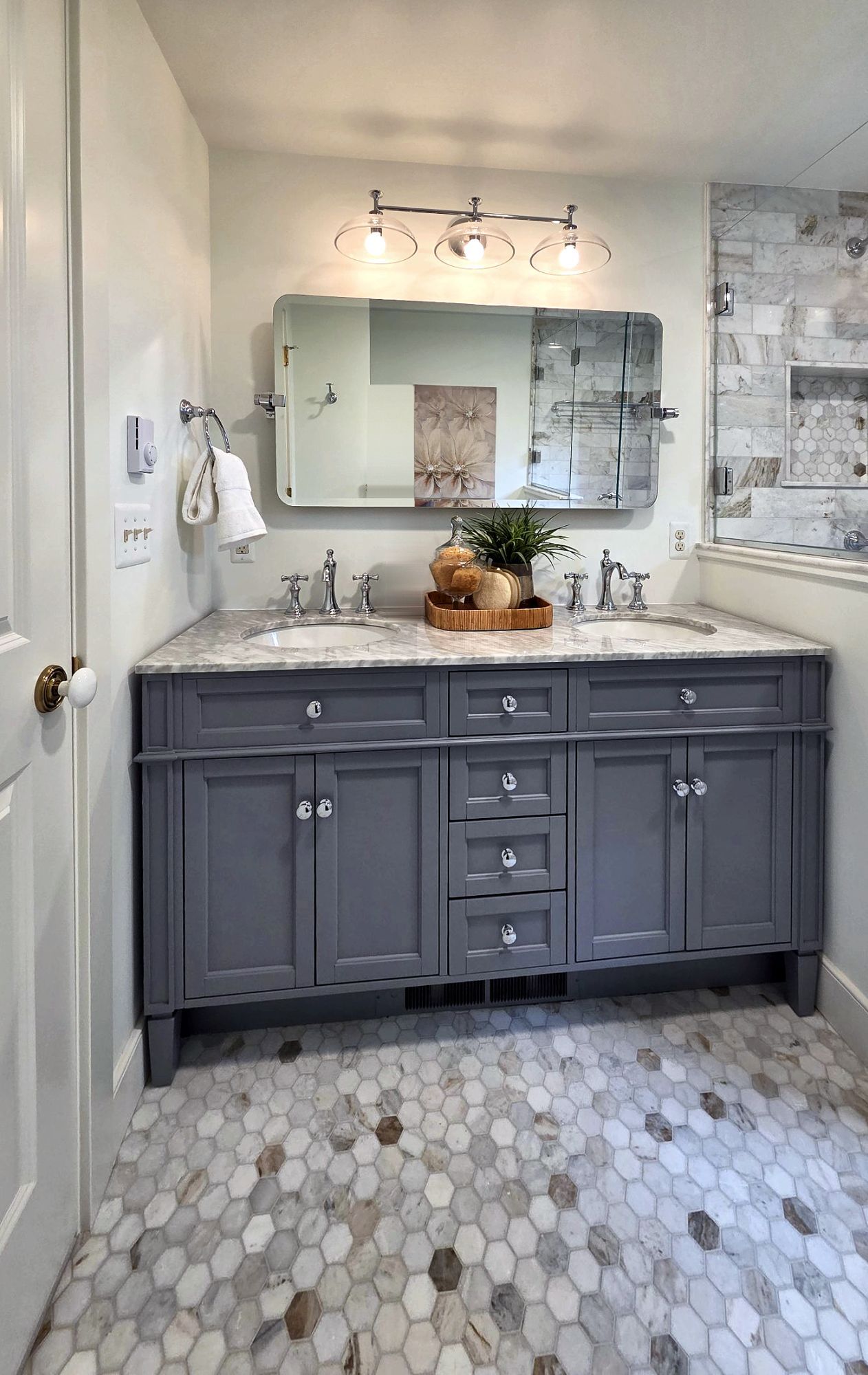Bathroom with a gray double vanity and marble top beside a glass-enclosed tile shower with a niche, over a marble hex tile floor.
