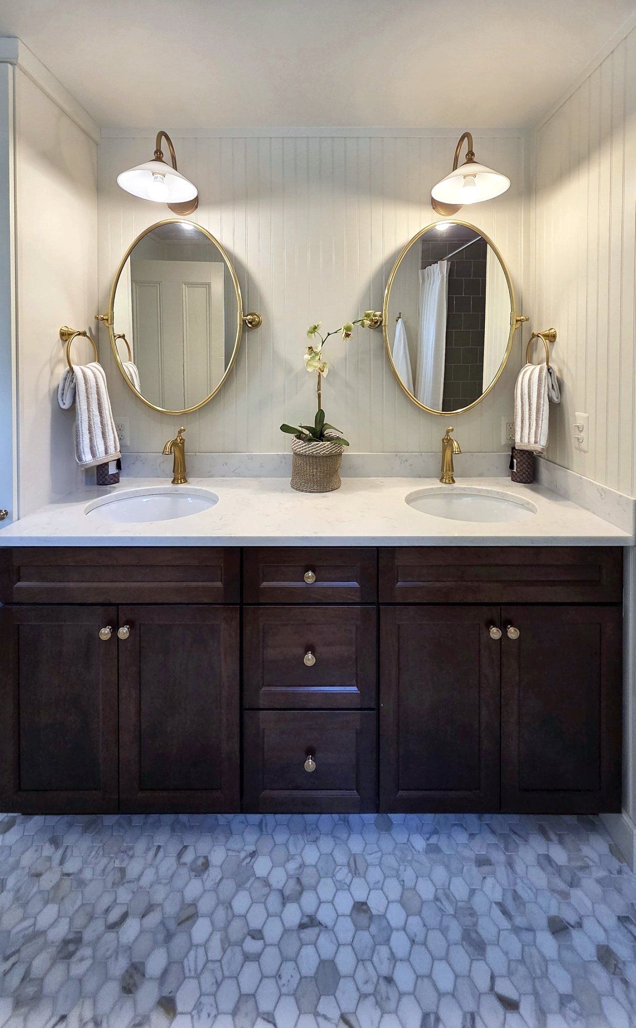 Bathroom with a dark stained double vanity, white quartz top, brass faucets, oval brass mirrors, and beadboard walls.