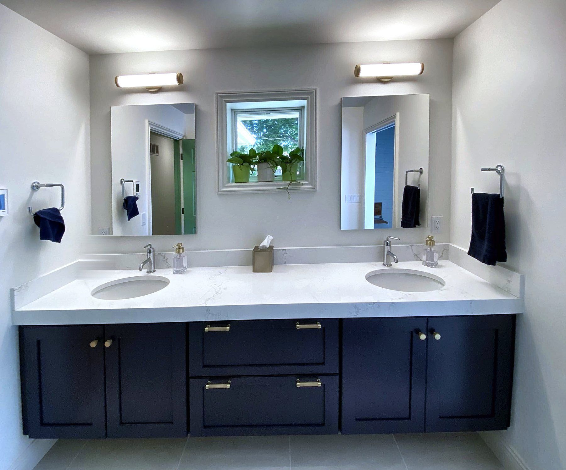 Primary bath with a navy floating double vanity, white quartz top, brass hardware, and a window between two mirrors.