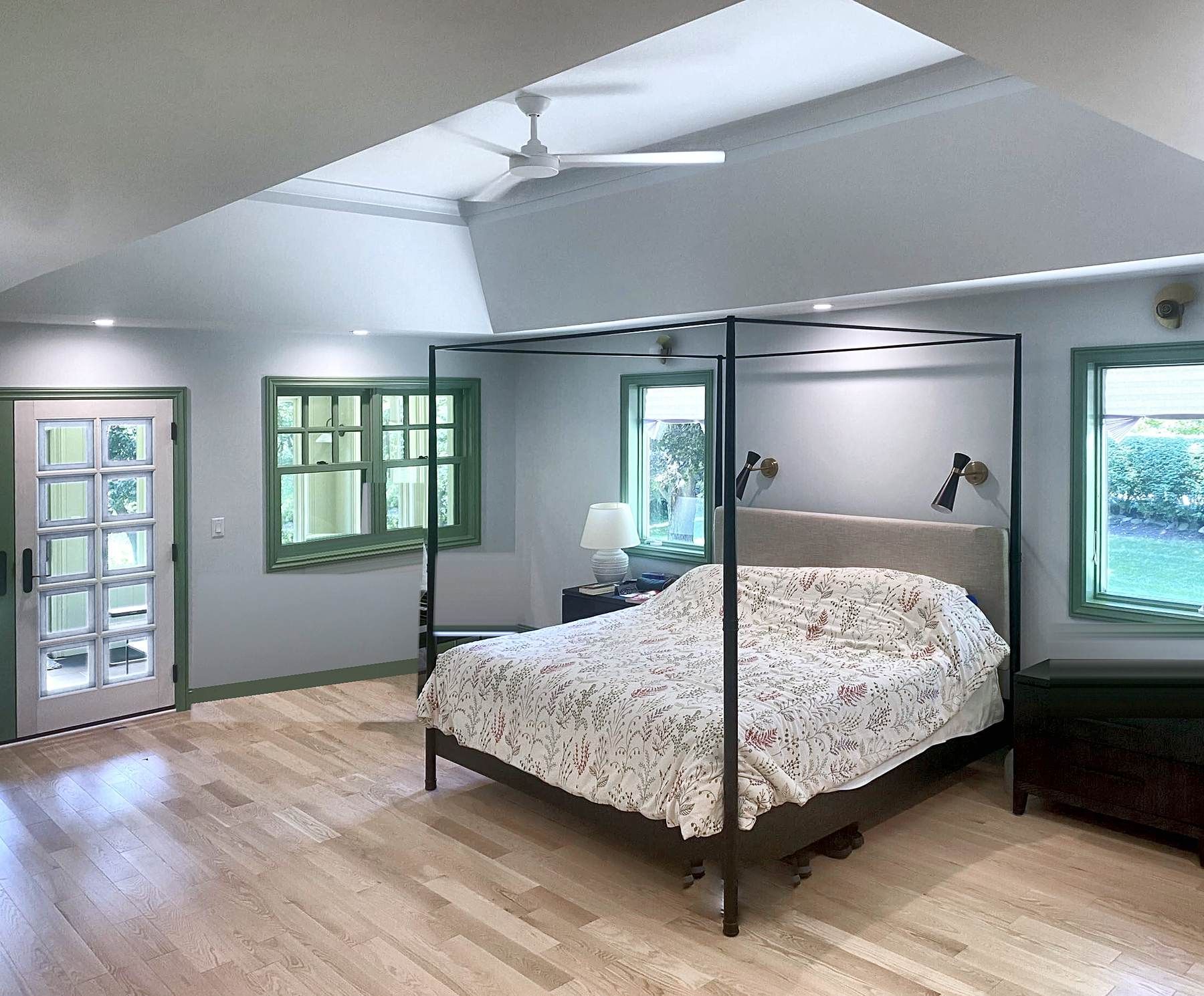 Primary bedroom in the addition with white oak floors, a tray ceiling, green-trimmed windows, interior windows into the sun porch, and a glass door to the porch.