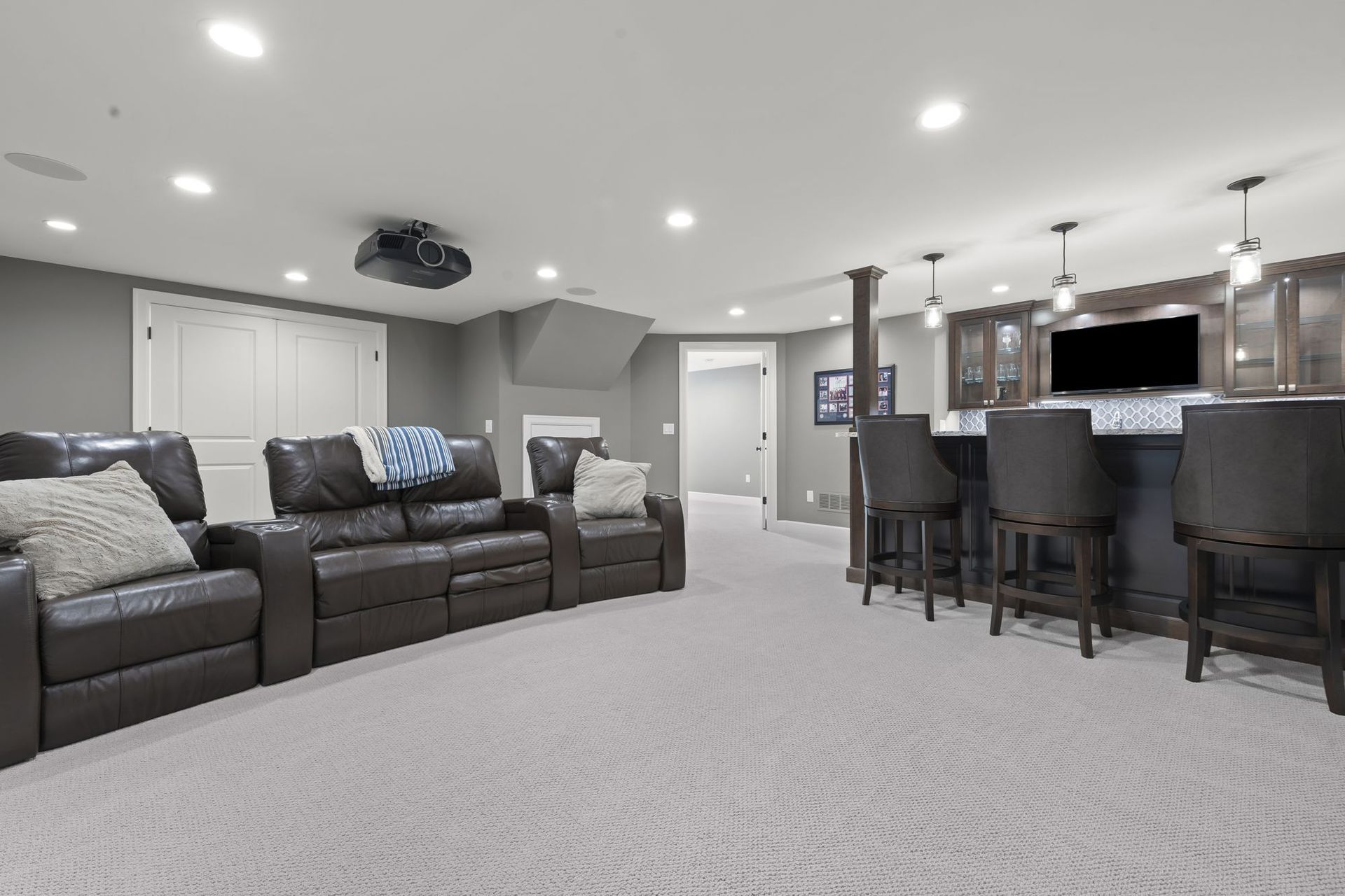 Finished walkout basement with theater seating, a ceiling projector, and a wet bar with leather stools.