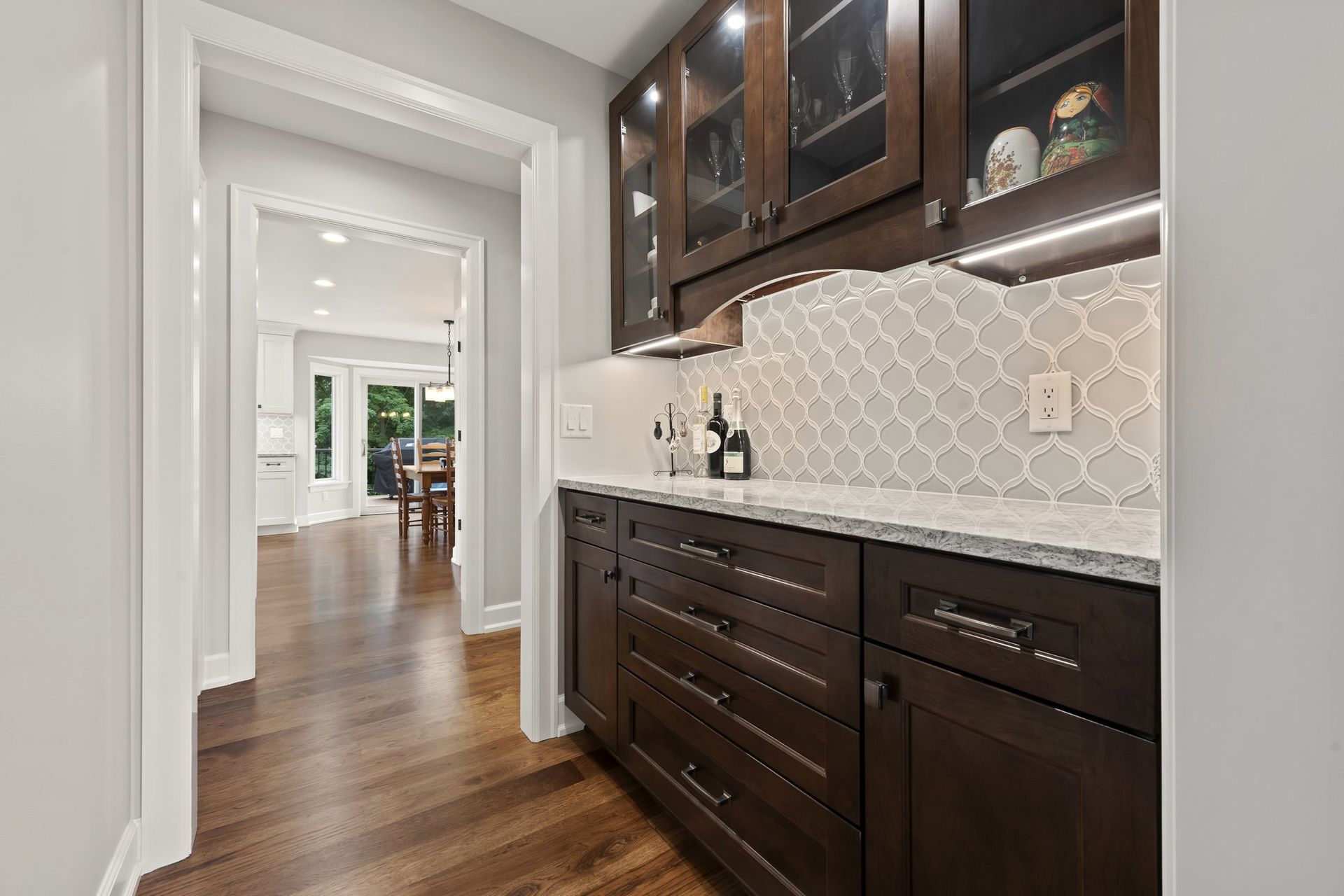 Butler's pantry with dark stained cabinets, glass upper doors, under-cabinet lighting, and a gray arabesque tile backsplash.