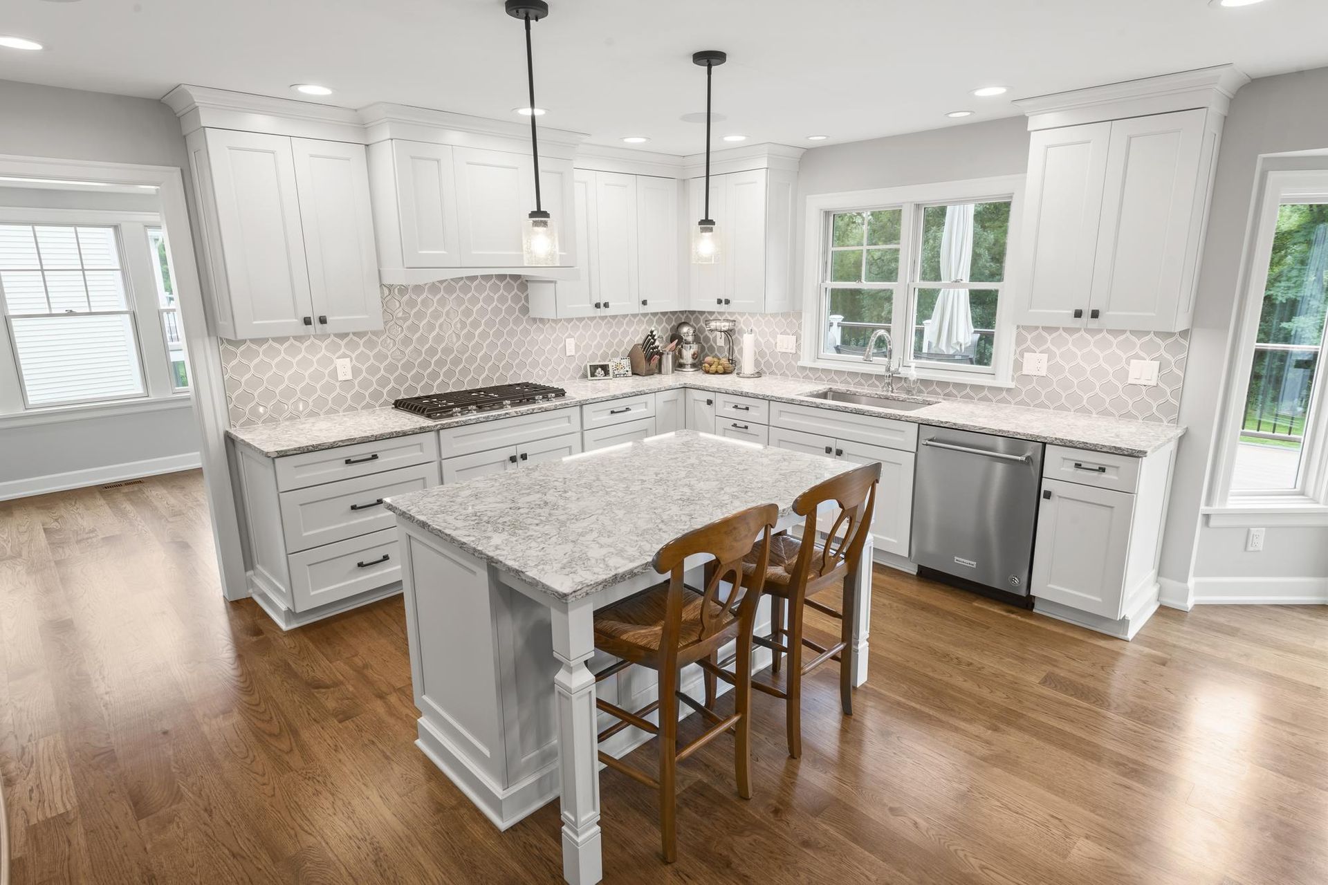 Remodeled kitchen with white shaker cabinets, a white island with two counter stools, a gray arabesque tile backsplash, and hardwood floors.